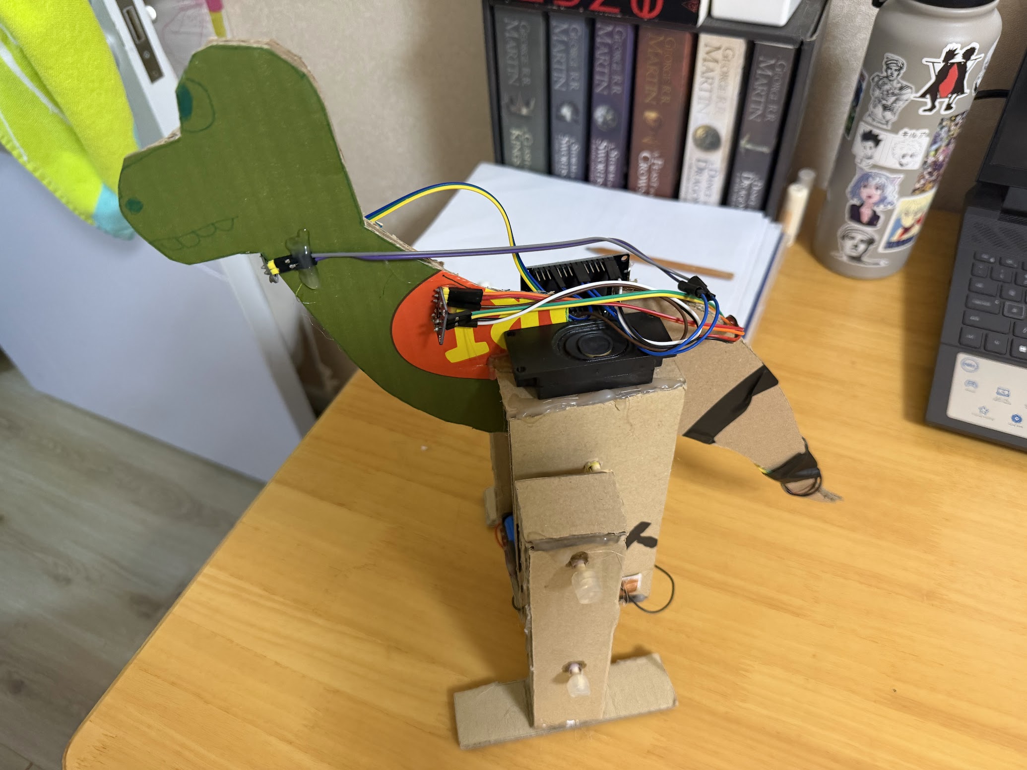 A cardboard dinosaur with electronics wired into its body, standing on a desk